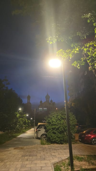 A nighttime street scene in Razvilka, Russia, captures a modern urban environment illuminated by a bright streetlamp. The foreground features a paved walkway with a tiled surface, bordered by grass and greenery. To the right, a parked silver SUV is partially obscured by dense foliage. Further down the walkway, a red car is visible. In the background, a church with multiple onion domes and crosses stands against a dark blue sky, suggesting it is either twilight or a heavily overcast night. The church is adorned with a row of illuminated square windows near its base. Surrounding the church and walkway are trees and residential buildings, contributing to the urban landscape. The streetlamp in the foreground casts a strong, warm light, creating sunburst-like rays and illuminating the leaves of a nearby tree. The overall atmosphere is one of a quiet, residential area at night.
