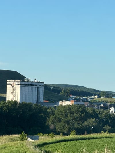 This image depicts a rural landscape in Pervomaiskoe, Russia. The foreground features a vibrant green field of crops, with tall grasses and a small, weathered concrete structure visible to the left. Beyond the field, a dense forest of dark green trees occupies the middle ground, partially obscuring a small cluster of buildings, possibly a village, nestled amongst rolling hills. In the background, a large, light-colored, multi-story industrial building with a prominent external staircase dominates the left side of the frame, set against a backdrop of a green, treed hillside. The sky is a clear, bright blue with no visible clouds.