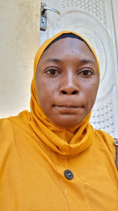 A frontal portrait displays an adult female with dark skin, wearing a mustard yellow hijab and a matching top with a black button on the front. A small silver stud piercing is visible on her left nostril. Her expression is neutral, and her gaze is directed forward. The background features a light-colored wall on the left side and a white door with embossed circular and linear decorative patterns, along with a silver-colored handle and lock mechanism, on the right. The scene depicts an indoor environment in the city of Madobi, Nigeria.