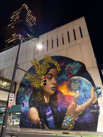 A vibrant mural dominates the scene, portraying a woman with a floral crown, planets, and hands holding a globe, set against a galaxy-like background. A tall building with illuminated windows stands in the background against the night sky. A traffic light showing red is positioned in front of the mural. A street sign indicates "one way".