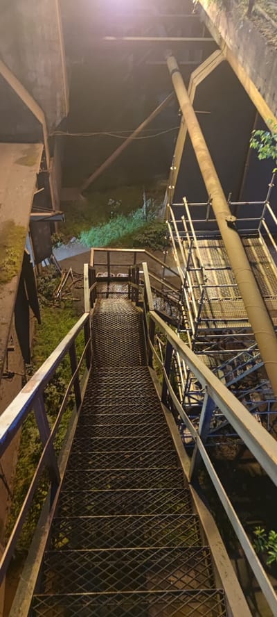 An outdoor, elevated metal staircase descends. The staircase is made of diamond plate metal treads and has metal railings on both sides. The area surrounding the staircase appears to be an industrial or construction site at night, with some overgrown vegetation visible on the ground below. There is a large pipe running vertically alongside the staircase and what appears to be scaffolding. The scene is lit by artificial lights, casting shadows and highlighting the textures of the metal and vegetation.