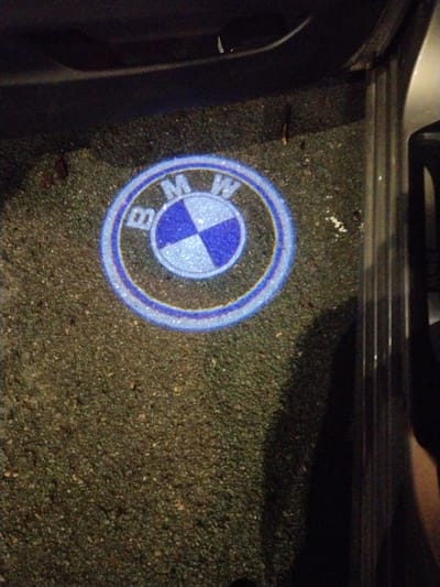 The image shows a BMW logo projected onto the ground, likely from a car's door light. The logo is circular with blue and white quadrants and the letters "BMW" visible. It is illuminated, appearing to be a light-up emblem. The ground beneath it is dark and textured, resembling asphalt or a gravel surface, with some small debris scattered around. To the right of the logo, a dark, vertical structure with a ribbed texture is visible, suggesting the side of a car door or trim. The overall lighting suggests it is either nighttime or a dimly lit indoor space. This scene is not located in the city of Častolovice, Czechia; the image depicts a car accessory and its installation.