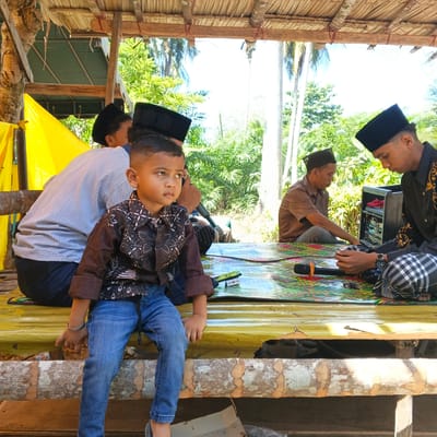 In Aluekeutapang, Indonesia, a group of people are gathered on a raised wooden platform, partially covered by a thatched roof. In the foreground, a young boy, wearing a patterned brown shirt and blue jeans, sits facing left, looking away from the camera with a contemplative expression. He wears a silver bracelet on his left wrist. To his left, partially visible, is a man in a light-colored, striped long-sleeved shirt and dark pants, also wearing a black peci (cap). Behind the boy, another man in a brown short-sleeved shirt and grey pants is seated, looking down at something in front of him, possibly interacting with a sound system. To the right, a young man in a black long-sleeved shirt with gold embellishments and a black peci is seated, looking intently at his hands, which are resting on a small electronic device, possibly a phone. He wears checkered pants. A black speaker box with red fabric inside is positioned to his right. The platform is covered with a colorful, patterned mat, on which rests a black microphone with an orange pop filter. A packet of "Lucky Strike" cigarettes is also visible on the mat. The background reveals lush green vegetation and palm trees, indicating a tropical environment. A bright yellow tarp is visible on the far left, attached to a wooden support. The overall scene suggests an informal gathering or event taking place outdoors.