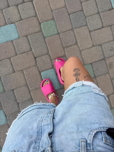 The image captures a top-down view of a person's legs and feet on a paved surface, likely an outdoor path or sidewalk in Huntington Beach, USA. The person is wearing light blue denim shorts with a distressed, frayed hem. On their right upper thigh, several outline tattoos are visible, including a seashell, a flower (possibly a hibiscus), and another shell or flower bud, along with a less defined tattoo higher up the leg.
The person wears bright pink slide sandals, and their toenails are painted a vibrant blue. A white beaded anklet adorns the right ankle. The ground is composed of rectangular pavers laid in an alternating pattern, predominantly in shades of grey and brown, with some pavers showing faint greenish or bluish hues. The scene conveys a casual, summery atmosphere. No specific time of day or weather conditions are explicitly visible. There is no discernible text in the image.