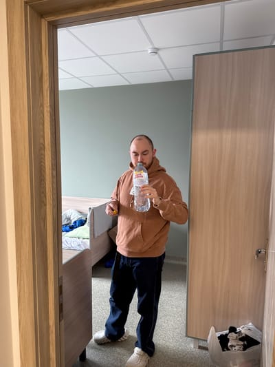 The image, when correctly oriented, shows a young man drinking from a water bottle inside what appears to be an institutional or commercial building, consistent with a location such as Borovoye Matyushino, Russia. The man, likely in his 20s or early 30s, has short hair and a beard/stubble. He is dressed in a light brown hoodie, dark blue jeans, and white athletic-style shoes. He holds a clear plastic water bottle, clearly labeled "SAINT SPRING," to his mouth, actively taking a drink.
He stands next to a light-colored wooden desk or table. On the desk, partially obscured, are some folded clothes in blue and white. The room features light green/grey walls and a white suspended ceiling with a grid pattern, from which a white smoke detector protrudes. The floor is covered with a light grey carpet. In the background, to the man's right, a white plastic bag containing dark clothes hangs or rests on a surface. Light wooden door frames are visible in the foreground (left) and background (right), with a door handle and lock plate visible on the background door. The lighting is consistent with an indoor setting, and the man's expression is neutral, focused on drinking.