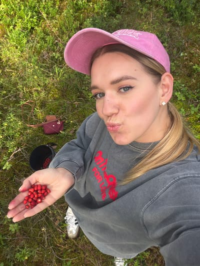A young Caucasian woman with blonde hair, wearing a pink baseball cap and a grey sweatshirt, is taking a selfie outdoors. She is holding a handful of small wild strawberries in her open palm. A black bucket, containing more strawberries, is visible to her left. Behind her, a small maroon bag with a brown flap is lying on the grassy ground. The environment appears to be a natural, grassy area with low-lying green vegetation and some patches of dirt. The background suggests a rural or park-like setting, potentially on the outskirts of a city. There is no specific indication of the city of Mucenieki, Latvia, in the image itself, but the scene is consistent with a natural landscape found in such a region.