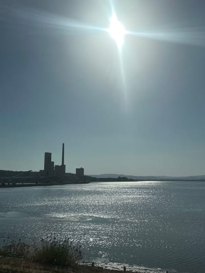 The image shows a body of water with a silhouetted industrial complex on the horizon. The sun is high in the sky, casting a bright glare on the water. The industrial complex appears to be a power plant or factory, with several tall structures including a smokestack. The water reflects the sunlight, creating a sparkling effect. The foreground shows a patch of dry vegetation along the shoreline. The scene is likely a waterfront industrial area. This image was captured in the city of Bages, France.
