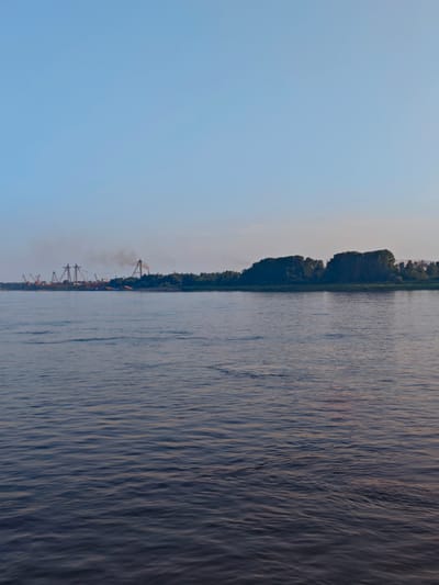 A wide, calm river occupies the foreground of the image. The water is a deep blue-grey, with gentle ripples reflecting the clear, pale blue sky above. On the far bank, a dense line of dark green trees forms a natural horizon. Beyond the trees, industrial structures are visible, including tall cranes and what appear to be loading facilities. Smoke billows from one of the structures, suggesting ongoing industrial activity. The scene is likely set during the early morning or late evening, given the soft light and clear sky. The location appears to be the Angara River in Lesosibirsk, Russia, a city known for its timber and pulp industries, which would explain the presence of industrial infrastructure on the riverbank. There are no people or animals visible in the image.