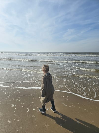 The image captures a person standing on a sandy beach at the water's edge, gazing out at the sea under a clear sky. The individual, who appears to be a young person with light-colored hair, is dressed in a muted-toned coat, dark trousers, and sneakers. They are positioned on the wet sand, where foamy white lines from gentle waves are receding.

The ocean stretches into the distance on the left, displaying moderate chop and whitecaps along the shoreline. Above the horizon, the sky is a pale blue, adorned with delicate, wispy clouds. To the person's right, a distinct, elongated shadow extends across the sand, suggesting the sun is at a moderate angle, indicative of either mid-morning or late afternoon. The scene conveys a tranquil moment by the coast, likely in Ostend, Belgium. No text is visible in the image.