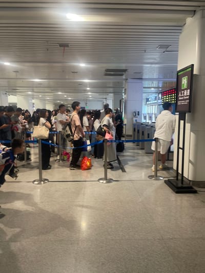 The image depicts a queue of people waiting to pass through a security checkpoint inside the Jinhua Railway Station in Jinhua, China, located at latitude 29.381 and longitude 120.04. The queue is lined up along a blue rope barrier, leading towards a security gate marked with a sign reading "人工通道" (Manual Channel) in Chinese. The people in the queue are mostly adults, with a few children, and they are dressed in everyday clothing. They are standing patiently in line, with some looking at their phones or around the station. The station's interior is characterized by a white tiled floor, a high ceiling with white panels, and overhead lighting. The scene suggests a busy travel environment, with many people waiting to pass through security before boarding their trains.