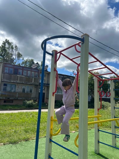 A young girl is climbing on a playground monkey bar structure. She is wearing a light purple tracksuit and sneakers. The playground equipment is made of metal and wood, with blue, red, and yellow accents. In the background, there is a building with several windows and balconies, a green grassy area, and trees. The sky is partly cloudy. The location is identified as Malaya Borshchevka, Russia.