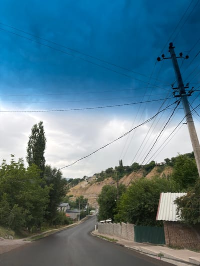 The image depicts a street scene in Khumsan, Uzbekistan. A paved road curves into the distance, flanked by green trees and vegetation on the left and a brick wall with a corrugated metal roof structure on the right. In the background, a steep, dry hillside is dotted with scattered houses and trees. A prominent utility pole with numerous electrical wires is situated on the right side of the road, extending upwards into a bright blue sky with scattered clouds. The overall impression is that of a semi-rural or suburban residential area in a hilly region.