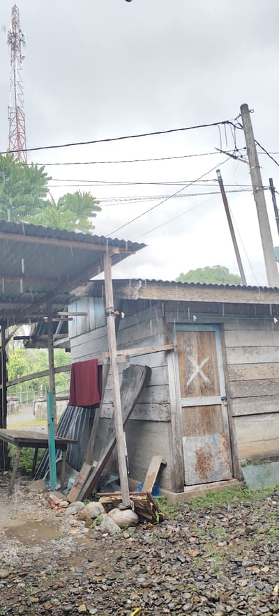 The image shows a rural scene in Jijiem, Indonesia, under a cloudy and rainy sky. In the foreground, a rough, cobbled ground is visible, wet from the rain, with scattered rocks and some green vegetation. To the left, a wooden table is partially visible. Behind the table, a section of a structure with a corrugated metal roof is present, also appearing wet from the rain.

The main subject of the image is a small, rustic wooden house or shed. The walls are constructed from weathered, grey-toned wooden planks. A wooden door with a blue frame and a white "X" painted on it is centrally located on the front of the structure. To the left of the door, a maroon-colored piece of fabric is hanging from a wooden support.

In the background, a tall communication tower with an orange and white lattice structure is visible against the overcast sky. Power lines are strung across the image, supported by wooden poles, one of which is to the right of the wooden structure. Trees with green foliage are present behind the structures. The overall impression is one of a simple, possibly impoverished, rural environment experiencing inclement weather.