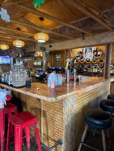 The interior of a bar, located in Hanalei, United States of America, is depicted. The establishment exhibits a Polynesian-themed design, characterized by bamboo ceiling beams, woven matting on the ceiling and walls, and hanging white and green shamrock cutouts. A central bar counter, clad in woven matting with a light-colored, patterned top, extends through the scene. Two red metal bar stools are visible on the left, and multiple black upholstered round stools with wooden legs occupy the right. The bar is equipped with a large clear plastic self-serve water dispenser labeled "SELF SERVE WATER STATION," silver pitchers, and a television displaying "ALOHA" on the left side. Bottles of various liquors, glassware, and point-of-sale tablets are arranged behind the counter. A black cylindrical object, a lime wedge, and stacked clear cups draped with a white cloth are on the counter surface. The right side of the bar-back features a prominent framed portrait flanked by numerous smaller framed photographs, all adorned with yellow floral garlands and white candles, forming a memorial or decorative display. Two draft beer taps are also on this section of the counter. No individuals are present within the scene.