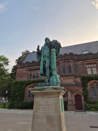 A full-shot, low-angle, outdoor photograph captures a weathered bronze statue of John Witherspoon (1723-1794) situated on a stone pedestal in front of a large, Gothic-style stone building with ivy covering portions of its exterior. The statue depicts Witherspoon in period clothing, with his right arm extended upward and his left hand resting on a globe supported by a column. The building behind the statue features arched windows and intricate stone carvings. The sky above is a pale blue with scattered clouds. The ground in front of the statue is paved with interlocking bricks and some concrete. The image appears to have been taken in Princeton, New Jersey, USA, likely on the campus of Princeton University, given the architecture and the presence of the statue of John Witherspoon, a prominent figure in the university's history.