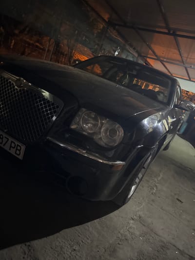 A dark-colored Chrysler 300C sedan is parked in a dimly lit, covered outdoor structure, likely a carport or garage, in Krushare, Bulgaria. The vehicle is angled towards the viewer, showcasing its front left quarter. Its large, chrome-accented grille with a mesh pattern is prominent, topped by the Chrysler emblem. Dual circular headlights with clear lenses are visible on the front left. A partial license plate, displaying "87 PB," is affixed to the lower left of the front bumper. The ground beneath the car is rough and uneven, appearing to be concrete or compacted dirt with scattered debris. Overhead, a metallic frame structure supports a translucent or reflective roof material, from which warm, orange-toned light reflects onto the car's surface and windshield. No individuals are present in the image.