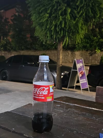 The image shows a plastic bottle of Coca-Cola standing on a wooden table. The bottle is partially filled with a dark liquid, presumably Coca-Cola. In the background, there are cars parked on the street, a tree with green foliage, and a purple A-frame sign advertising Milka chocolate. The scene appears to be outdoors at night, possibly in a city. The overall impression is that of a casual setting, perhaps a cafe or a roadside stop. The location is identified as Biaga, Bulgaria.