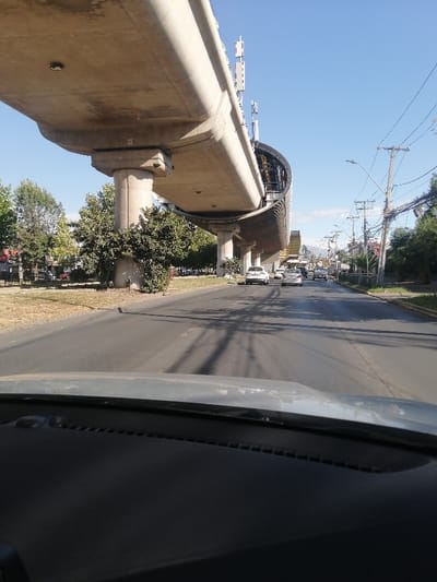 Captured from inside a vehicle, the image showcases a road scene under a sunny sky. An elevated railway structure, possibly for a metro or train, dominates the left side, with pillars supporting its concrete track. Several cars drive along the road. Trees and foliage line the sides. Utility poles with power lines run alongside the road. In the background, distant mountains are visible against the clear sky. The dashboard of the vehicle occupies the lower portion of the frame.