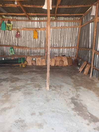 The image depicts the interior of a basic, utilitarian structure located in Zangon Aya, Nigeria. The structure features walls constructed from corrugated metal sheets fixed to a visible framework of rough-hewn wooden beams. A primary wooden post stands vertically in the center-left foreground, extending from the concrete floor to the overhead wooden roof supports. The floor is a stained and uneven concrete slab.
Illumination is provided by a single bare incandescent light bulb hanging from the roof structure near the upper left corner, supplemented by natural light entering from an unseen opening.
Hanging from various horizontal wooden beams are several items: two green fabric tote bags (one with partial "MELON" text visible) are on the upper left, a reddish-brown patterned fabric item is centrally located, and a yellow fabric item is further right. Below these, a green mesh bag hangs on the left. Two white fabric pieces are draped over a beam above the central wooden post.
Against the left wall, a dark, rolled-up mat or tarpaulin rests on the floor, next to a stack of green items, possibly containers. Along the rear corrugated metal wall, numerous rectangular wooden panels or boards are leaned vertically against the surface, some exhibiting faint markings or perforations. To the far right, another smaller stack of similar wooden pieces is present.
No individuals, actions, or direct interactions are visible within the scene.