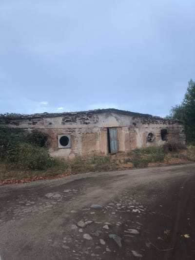 The image shows an abandoned, dilapidated brick building with a damaged roof. The building has several openings, including a doorway with blue wooden doors and at least two circular vents or fans mounted on the exterior walls. Overgrown vegetation surrounds the building, and a dirt road with potholes is in the foreground. The sky is overcast. The scene is located in Нуъмон Розиқ, Tajikistan.