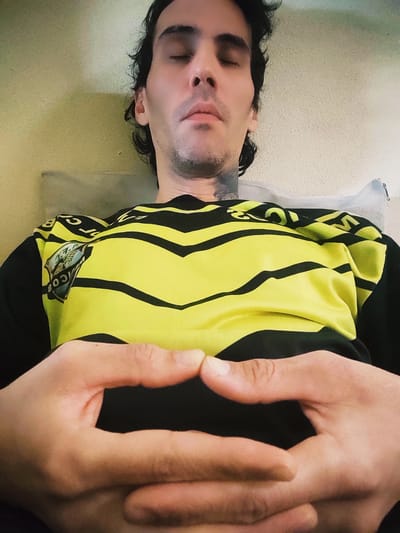 A man is lying down on a bed or similar surface. He is wearing a yellow and black shirt. His eyes are closed, and his hands are resting on his stomach. The image was likely taken indoors. There is no indication that this image was taken in Remedios de Escalada, Argentina.