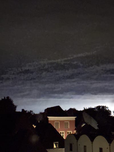 The image shows a nighttime view of buildings in Deventer, Netherlands, with a dark, cloudy sky overhead. A prominent pink building with white-framed windows is visible in the center, illuminated from within. Dark silhouettes of trees and other rooftops surround it. The sky has a layered, textured appearance with areas of lighter and darker clouds, suggesting a stormy or overcast atmosphere.