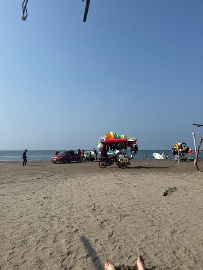 The image displays a daytime beach scene located in Antón Lizardo, Mexico. The sky is clear and cloudless, exhibiting a light blue hue. In the immediate foreground, two bare human feet are visible, positioned on light brown sand marked with numerous footprints.
In the mid-ground, a sandy beach extends towards the ocean. A lone individual, dressed in dark clothing, is observed walking leftward across the sand. Closer to the water's edge, a red pickup truck is parked on the sand. Multiple people are gathered around the truck and along the waterline.
To the right of the truck, a mobile vendor cart is visible, constructed from a motorcycle with an attached wheeled trailer. This cart features a red canopy and is laden with numerous colorful inflatable rings on top, with various garments and bottled items hanging below. Further to the right, another vendor stand, also displaying colorful merchandise, is positioned closer to the ocean.
In the background, the calm blue ocean meets the horizon. A small white boat with an outboard motor is present in the water, to the right. At least one human figure is discernible wading or swimming in the ocean. Dark, linear elements and a rope-like object are partially visible in the upper corners of the frame, indicating overhead structures.