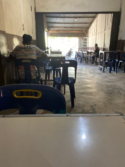 The image depicts an outdoor seating area of a restaurant or cafe in Lamreung, Indonesia. Two individuals are visible. In the foreground, a young male wearing a colorful, striped t-shirt sits with his back to the camera, facing a table. Further into the space, an older male sits at a table, facing the camera and appearing to be eating or drinking. The environment is characterized by simple wooden tables and blue plastic chairs arranged on a concrete floor. A corrugated metal roof covers the seating area, with open sides allowing natural light to enter. A motorcycle is parked to the left. The overall impression is of a casual, functional establishment.