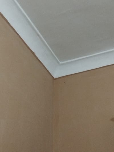 The image shows the corner of a room where the wall meets the ceiling. The walls are painted a light brown color, and the ceiling is white. A decorative white molding runs along the perimeter of the ceiling where it meets the walls. There are no people or other objects visible in the frame. The location is unknown, and there's no information in the image to indicate that the image was taken in Kuvasay, Uzbekistan.