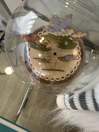 The image displays a clear glass globe containing a multi-tiered wooden diorama, located in Saint-Germain-en-Laye, France. The diorama features laser-cut and painted elements including a light blue butterfly with dark blue markings, several white daisy-like flowers with yellow centers, an orange-red multi-petaled flower, and various green foliage pieces. A string of small, warm-toned LED lights is visible within the wooden structure. The diorama's base is light-colored wood with a scalloped edge design. The glass globe exhibits reflections of blurred text, likely in French, and multiple bright, circular light sources. The surrounding environment includes a light brown wooden or laminate floor visible below and to the left of the globe. A shiny, silver-colored cylindrical object, likely a furniture leg, is also present in the lower left. To the lower right, a white, shaggy faux fur texture is partially visible, adjacent to a section of white and grey striped knitted fabric, indicative of a decorative gnome or plush item. A dark green or teal object is marginally visible at the bottom left edge. No individuals are present in the scene, and no actions or interactions are depicted.