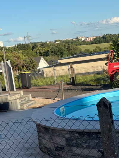 The image depicts an outdoor residential and semi-industrial landscape under bright daylight, situated in Pirmasens, Germany, with a vantage point looking northwest towards the commercial area around An der Ohmbach. No individuals are present in the scene.
In the immediate foreground, a dark gray chain-link fence spans the lower portion of the frame, with a rough-hewn concrete post vertically bisecting the right side. Behind the fence, a circular, above-ground swimming pool with a light blue cover is partially visible, indicating water beneath. The pool is integrated into a concrete-tiled patio area. To the left of the pool, concrete steps descend, showing wear and discoloration. A dark-colored pot rests on the patio to the left of the steps. The ground around the steps and behind the pool consists of dark grey gravel and reddish paving stones.
The midground features a residential backyard or garden area. A black wheeled refuse bin with a yellow lid stands upright near a metal fence. A dark hose runs across the gravel path. Behind the fence, a small section of a red compact loader or tractor-like machine, branded "WEIDEMANN," is partially visible on the right. Further back, a beige single-story building with dark-colored solar panels on its roof is prominent, flanked by greenery and another smaller white house with a dark roof.
The background reveals an expansive landscape dominated by a dense treeline, beyond which lies a large green field. In the far distance, several commercial or industrial buildings are visible. One notable structure is a white building with distinctive red and orange horizontal stripes. To its right, a building with a large yellow "M" logo, characteristic of a McDonald's restaurant, is identifiable. High-voltage power lines traverse the sky, supported by a tall lattice-style power pylon prominently centered in the upper background. The sky above is clear blue with scattered white cumulus clouds.