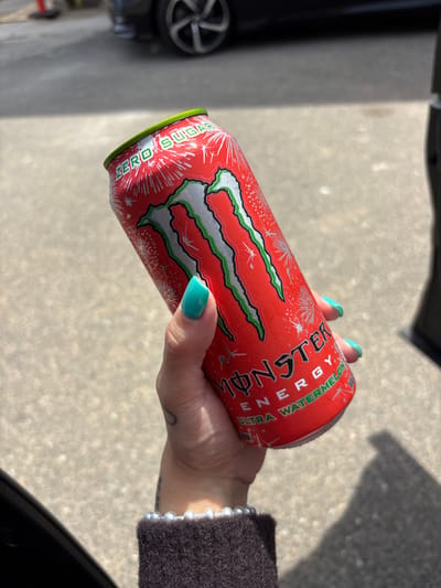 A person with turquoise manicured nails is holding a red and silver Monster Ultra Watermelon energy drink can. The can has the "ZERO SUGAR" label on the bottom. The person is wearing a dark, possibly knitted, sweater with a pearl bracelet. The background shows a concrete surface with shadows and a car wheel. This image was likely taken outside near a vehicle. There is no information in the image that directly points to Huntington Beach, United States of America. The inclusion of Huntington Beach would have to be extrapolated from other information.