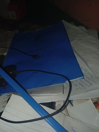 The image shows a messy desk with a blue folder, some papers, and electrical cords. The blue folder has some text and a date, "MAY 2020", printed on it. There are handwritten notes on some of the papers, including phrases like "Thank you for your" and "Valuable am exp". The environment appears to be an indoor setting, likely a room or office, with dim lighting. There are no people visible in the image. The scene does not provide any specific clues to determine its location in the city of Zongoma Kyari, Nigeria.