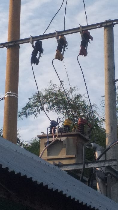 The image displays an overhead electrical utility installation in Lamreung, Indonesia, observed from a low-angle perspective. The scene features two concrete utility poles supporting a horizontal metal crossarm. Three fuse cutouts, colored dark grey, yellow, and red, are suspended from the crossarm. Below these, a beige-grey electrical distribution transformer is mounted, equipped with three top-mounted insulators colored blue, yellow, and red, to which primary electrical cables from the fuse cutouts are connected. Additional cables, some appearing light-colored, are routed to and from the transformer. Behind the electrical components, a dense cluster of green bamboo foliage is visible. In the lower foreground, a grey, corrugated metal roof of a building with visible fasteners occupies the bottom portion of the frame. A white PVC pipe, bent at an elbow, extends upwards from the roof area near the transformer. The sky above is partly cloudy. No people are present in the image.