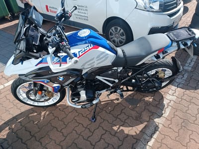 The image shows a white and blue BMW R1250 GS motorcycle parked on a paver brick surface. The motorcycle has a black seat and a small black bag attached to the rear. A white car is parked behind the motorcycle. Text on the car reads "Handwerker 2.0," "Werkstrasse 3," "67354 Römerberg," and a phone number. This indicates the location is likely Römerberg, Germany, a district of Germersheim. The scene is outdoors, with shadows cast on the ground, suggesting a sunny day. No people are clearly visible in the image.