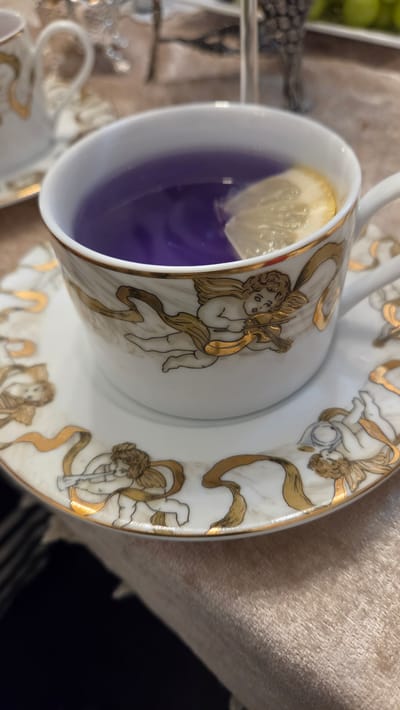 A close-up shot shows a white ceramic teacup and saucer with gold cherub designs and ribbons. The teacup contains a vibrant purple liquid, possibly tea, with a lemon slice floating in it. The saucer also features the same decorative elements. The scene is set on a textured, light-colored tablecloth. The background is blurred, suggesting an indoor setting, potentially a cafe or dining room in Keswick, Canada. No people are visible in the frame.