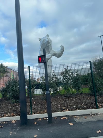 The image displays an outdoor urban scene in Argenteuil, France, under an overcast sky with patches of blue. The central subject is a large, white, polygonal sculpture of a bear, depicted standing upright on its hind legs with its left arm raised. In the immediate foreground, a dark grey pole supports a pedestrian signal displaying a red "don't walk" symbol, featuring two static human figures. To the left, another dark grey pole is partially visible. A green metal mesh fence runs horizontally across the midground, separating a paved dark asphalt surface with scattered brown leaves from a landscaped area. This landscaped section features low-lying green shrubs planted in dark earth covered with mulch and additional fallen brown leaves. Beyond the fence, various buildings, including what appears to be a multi-story structure with green architectural details, and sparse deciduous trees are visible in the background. A light grey concrete curb delineates the edge of the paved foreground.