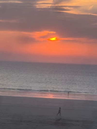 This image depicts a sunset over the sea at a beach in Pléneuf-Val-André, France. The sun, partially obscured by clouds, casts a vibrant orange and pink glow across the sky. The sea is calm, with a gentle reflection of the sunset on its surface. In the foreground, a sandy beach stretches towards the water. Two small figures are visible on the beach: one is running in the foreground, and another is standing further back near the water's edge. The scene suggests a peaceful evening at the coast.