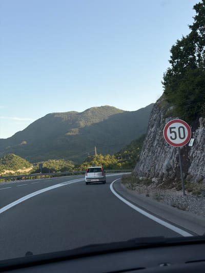 A white car is driving on a highway with a speed limit of 50 km/h. The highway is bordered by a rocky cliff face covered in trees on the right, and a forested mountain range on the left. The sky is clear and blue. The car's license plate reads "PG-EK023". The scene appears to be in Vrela, Montenegro.