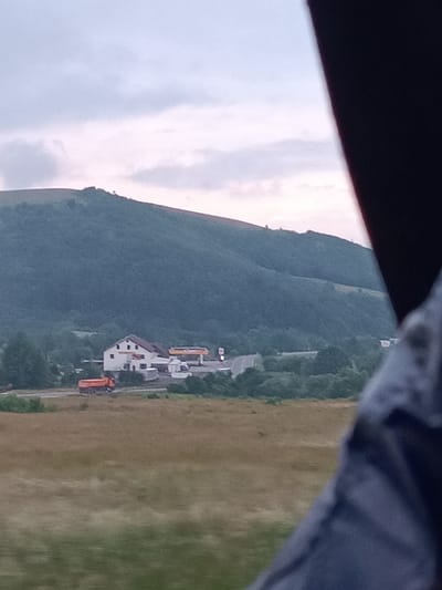An outdoor, slightly hazy, eye-level shot depicts a rural landscape with a focus on a building complex nestled at the base of a large, tree-covered hill. The sky is overcast with soft clouds. In the mid-ground, a white building with a red roof is visible, featuring several windows and what appears to be a smaller, attached structure. Adjacent to this building is a gas station with a yellow canopy and pumps. In the foreground, an orange dump truck is driving on a road in front of the building complex. The surrounding terrain consists of a grassy field in the immediate foreground and a dense forest covering the hillside in the background. The scene is set in Brețcu, Romania.