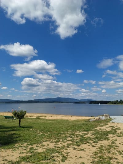 This is a landscape photograph taken on a sunny day at the Lipno Reservoir near Bližná, Czechia. The scene is dominated by a wide expanse of deep blue water under a bright blue sky with scattered white cumulus clouds. In the foreground, there is a sandy area transitioning to green grass. A small, sparse tree stands in the grassy area, with a green park bench situated to its left. On the beach, a few individuals are visible near the water's edge, and further out on the water, there are several small boats and what appears to be a person on a paddleboard. The background features a distant, hazy mountain range covered in dark green trees, with some lighter green foliage visible along the shoreline. The overall impression is of a serene, outdoor recreational setting.