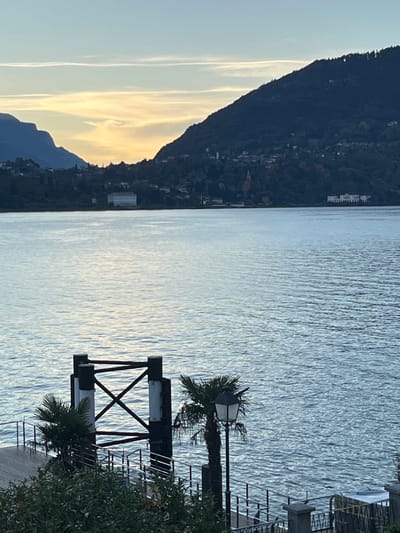 The photograph shows a lake scene with a dark, mountainous coastline on the left and calm, light-reflecting water in the center. On the right, there is a structure consisting of black beams and white platforms, with a palm tree growing from the top. A hint of architecture peeks over the mountain range. The sky exhibits shades of light blue and yellow, suggesting either sunrise or sunset.