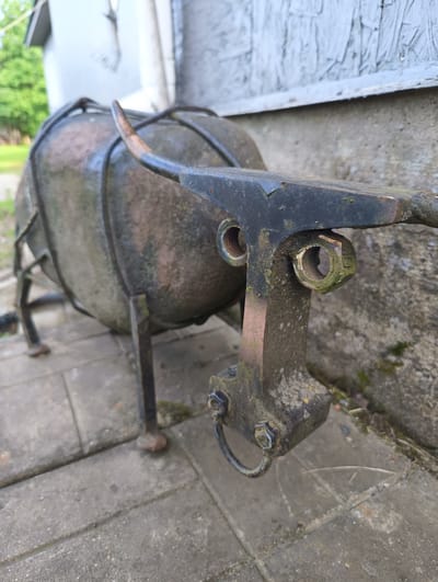 An outdoor, eye-level, close-up shot focuses on the head of a rusty, metal bull sculpture. The bull has a large, round body and appears to be constructed from salvaged metal parts. Its head is a dark, angular piece of metal with two visible bolts, one on each side. The bull's horn curves upwards and to the left. The sculpture is situated on a paved walkway, with a building's weathered siding visible in the background. The environment suggests a rural or semi-rural setting, possibly in or around Княжьи Горы, Russia.
