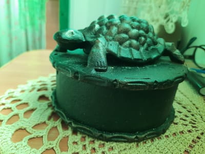 A dark, ornate, possibly cast-iron box sits on a crocheted doily. A decorative turtle figurine is attached to the lid of the box, its shell textured and its legs and head detailed. The box itself is dark green and hexagonal. The doily is a pale yellow-green color with an intricate openwork pattern. The background is blurred and appears to be an indoor setting, with hints of green curtains and a wooden table surface. The lighting casts a green hue over the entire scene. There are no people visible in the image. The scene is a still life arrangement of decorative objects.
