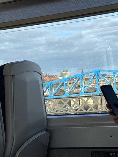 From a train window, the iconic blue framework of Tower Bridge dominates the view. In the background, a cityscape emerges, punctuated by a church spire. A hand holds a phone, partially obscuring the scene. The interior of the train is visible, featuring a light grey seat with a fold-down tray table. A sign indicating power outlets is on the wall.