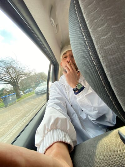 A male individual is positioned in the rear left passenger seat of a vehicle in La Wantzenau, France. He is wearing a white beanie, a white hooded jacket, and a nose ring. A watch with a green face, a dark strap, and a blue wristband is present on his left wrist. His right hand is raised to his mouth, holding a lit cigarette from which smoke is visible. The vehicle's interior features a light-colored headliner and a grey fabric seat on the right. Through the open window, a daylight street scene is visible, featuring a large deciduous tree with bare branches, a blue wheeled waste bin, and a light-colored hatchback with French license plates parked on the street. Additional residential buildings and vehicles are visible in the background.