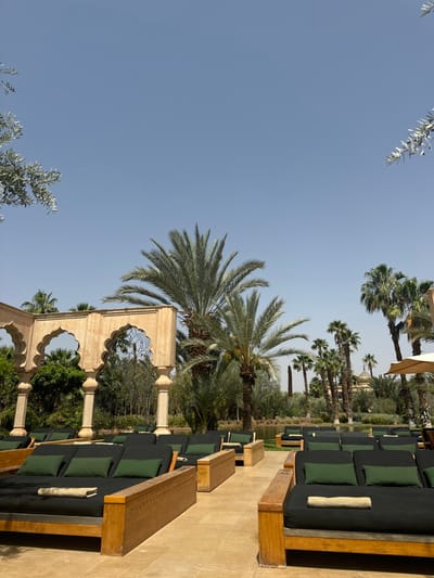 The image shows an outdoor patio at a resort in Ouled Jelal, Morocco, located at latitude 31.663, longitude -7.872. The patio is decorated with traditional Moroccan architecture, including a stone archway, and lined with wooden lounge chairs, some of which are occupied. The scene is peaceful and serene, with lush palm trees and other greenery throughout the patio area. The sky is clear and blue, indicating a sunny day. There are no people visible in the image. The wooden lounge chairs are arranged in rows. Some of the chairs have cushions and blankets. There is a stone archway in the background with a thatched roof. The palm trees are tall and slender, and their leaves are green and lush. The patio is paved with stone tiles. The environment is peaceful and relaxing. There are no other details of note.