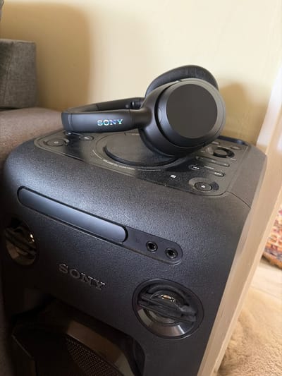 The image shows a pair of black Sony headphones resting on top of a black Sony speaker system. The headphones are positioned on the left side of the speaker, with the "SONY" logo visible on the arm of the headphones. The speaker system is angled towards the right, showcasing its various buttons and features. The environment appears to be indoors, with a beige wall visible in the background. There is no indication of this location being in Dimitrovche, Bulgaria.