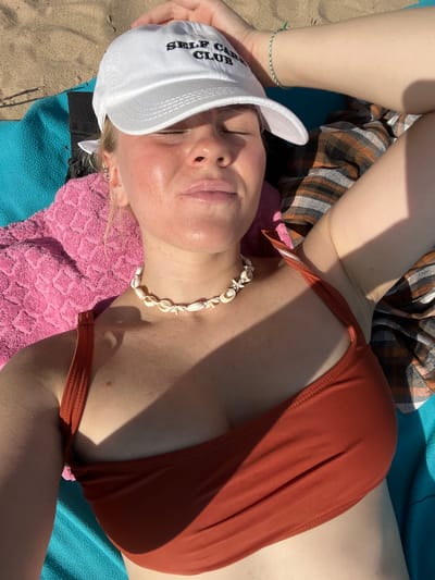 The image shows a young woman lying on a beach. She is wearing a white baseball cap, an orange bikini top, and a shell necklace. The woman is lying on a blue towel, with a pink towel and a plaid blanket underneath her. The background is a sandy beach with some vegetation. The lighting suggests it is a sunny day. The location in the city of Ust'ye, Russia is not discernible from the image.