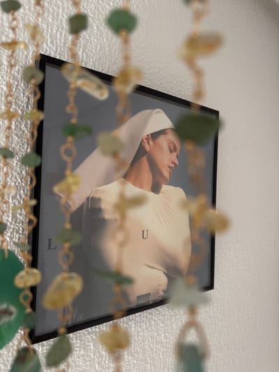 The image shows a framed album cover featuring a close-up of a woman with her eyes closed, resting her head on a cream-colored fabric. She is wearing a white headscarf that drapes over her shoulder. The album cover itself is framed in black and appears to be displayed against a textured white wall. In the foreground, out of focus, is a beaded strand with green and yellow beads, adding a decorative element to the scene. The album cover has text visible, including "PARENTAL ADVISORY EXPLICIT CONTENT" in the bottom right corner. Letters "U" and "L" are also visible on the album cover. The lighting suggests an indoor setting, possibly during the day. The overall mood is serene and artistic. The setting is identified as Las Rozas de Madrid, Spain.